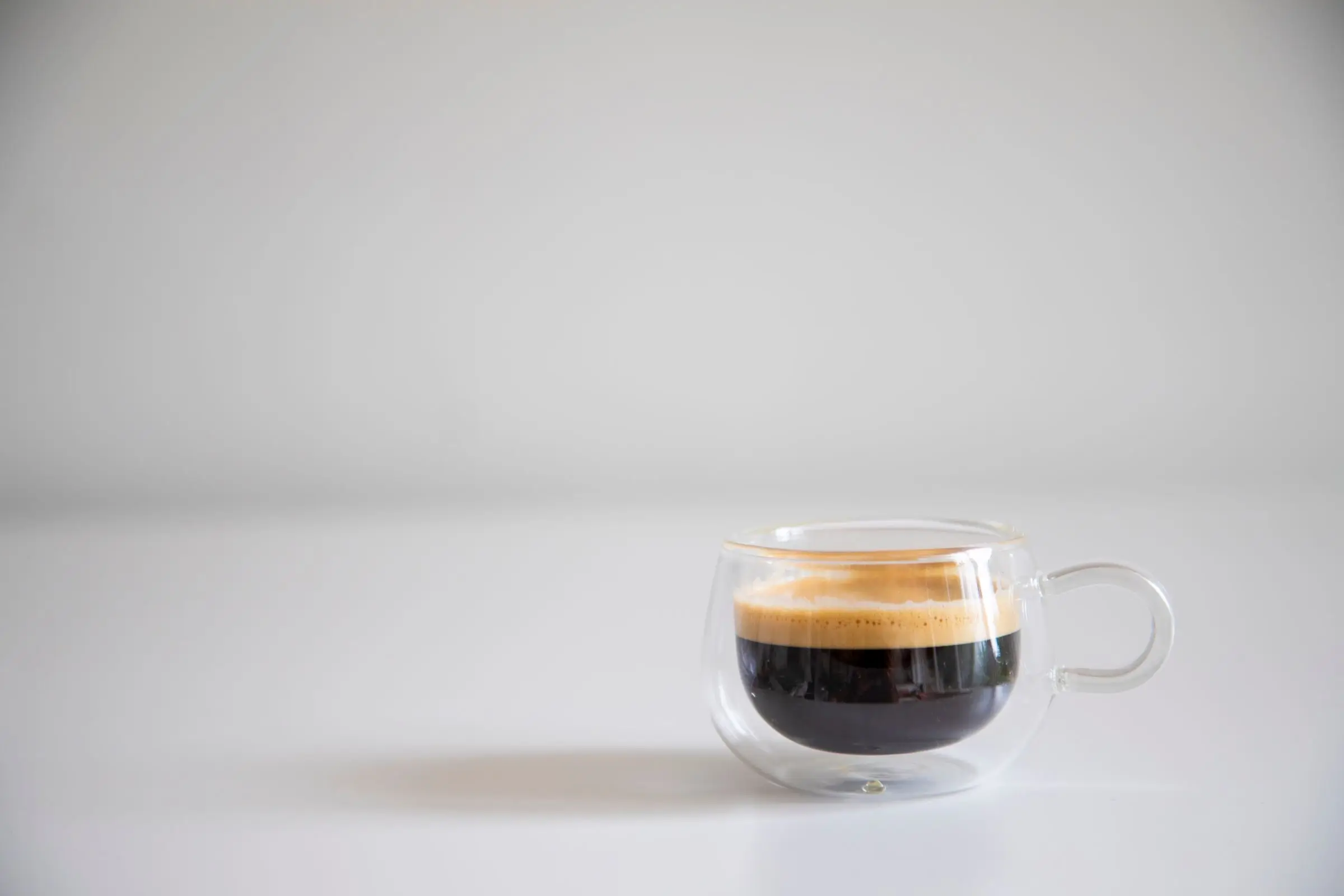 A double-walled glass cup of espresso with a pale crema, on a plain white surface with a soft shadow.