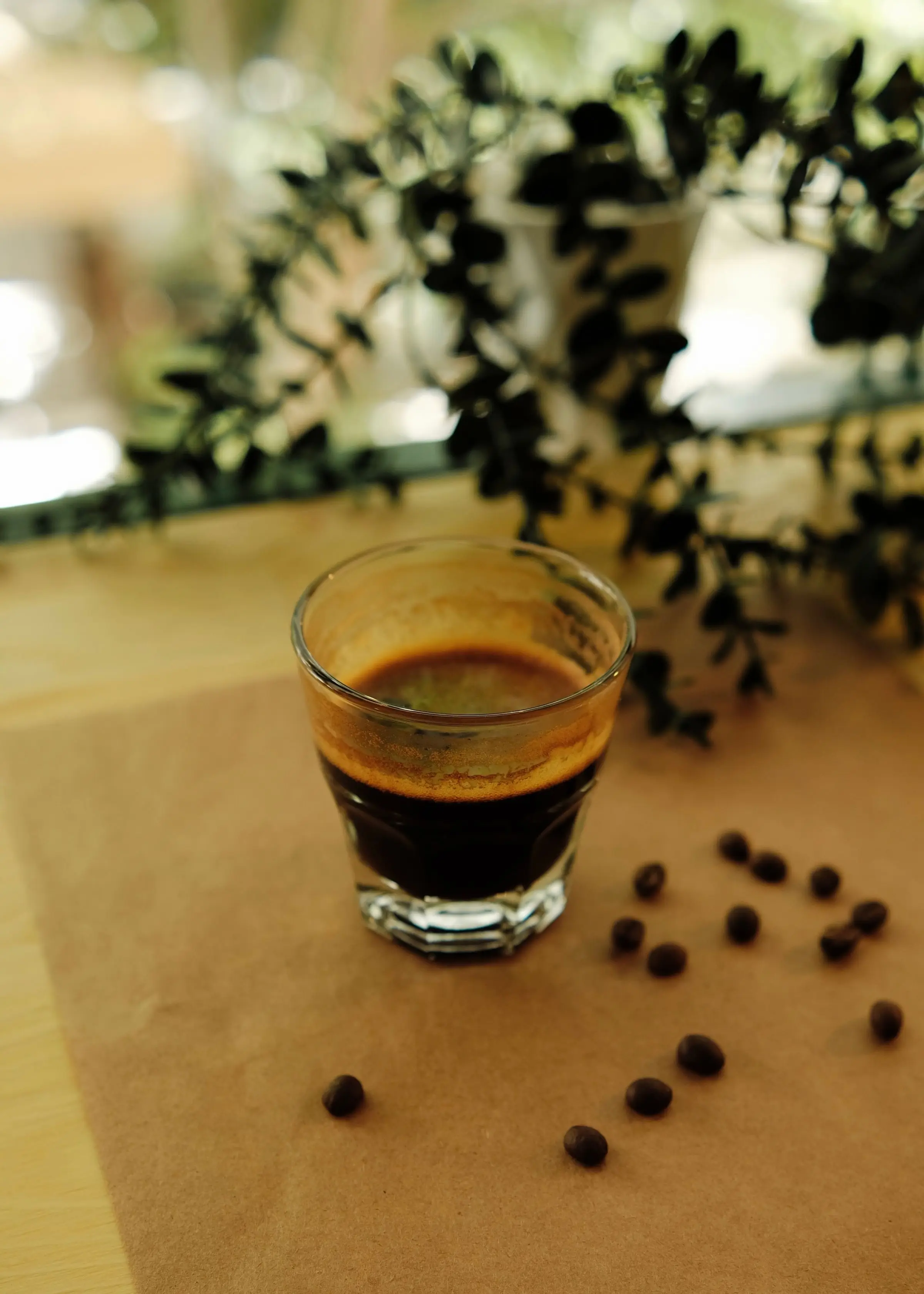 A small glass of espresso on brown paper with roasted coffee beans scattered around it.