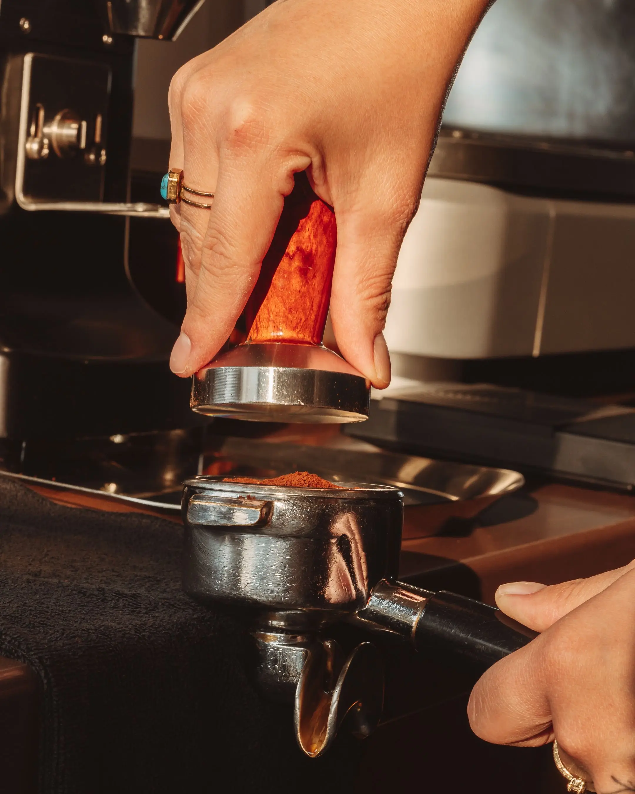 A hand pressing a wooden tamper into a portafilter of ground coffee in warm low sunlight.