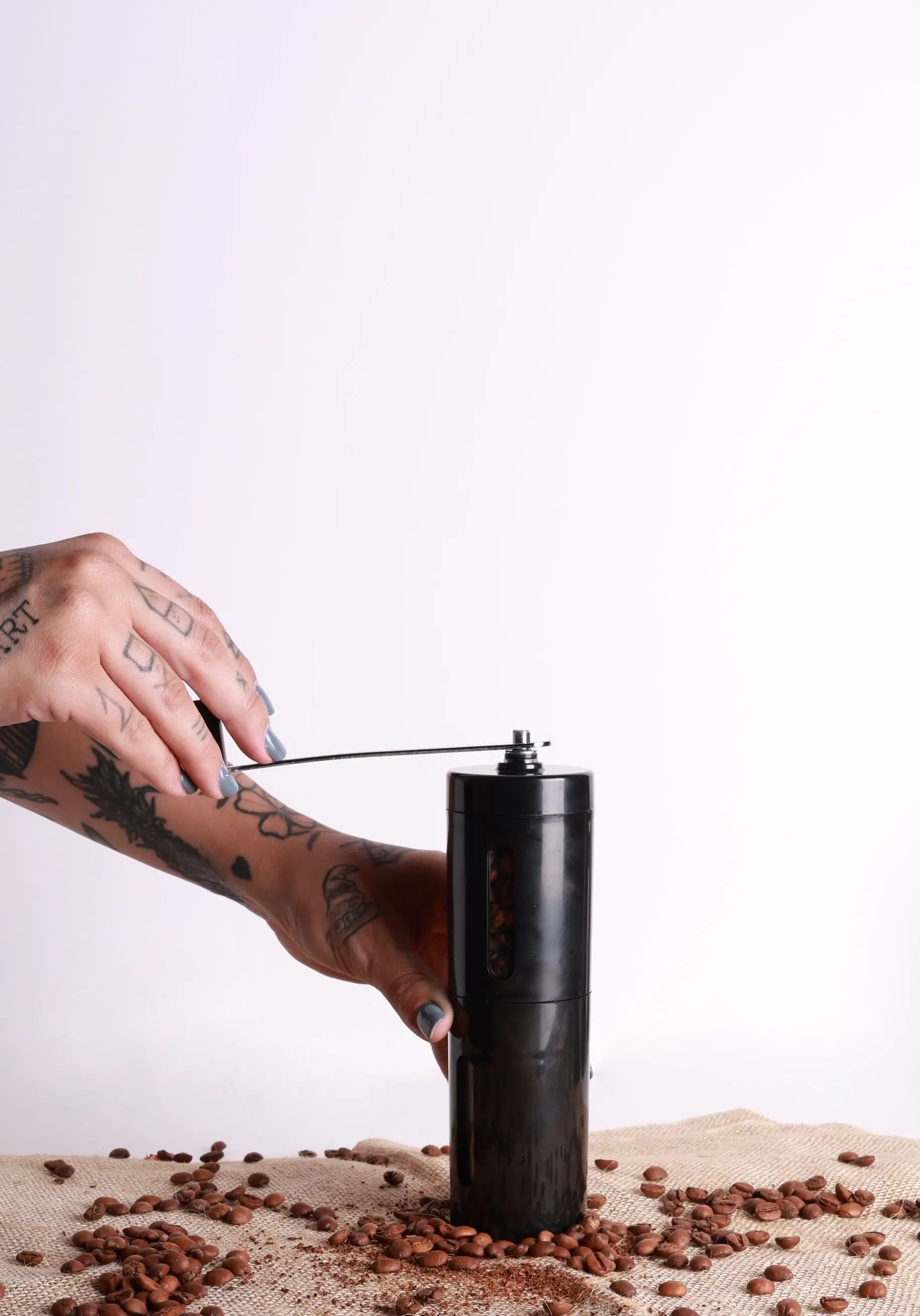 A tattooed hand turning the crank of a black hand grinder, with roasted beans scattered on hessian around it.