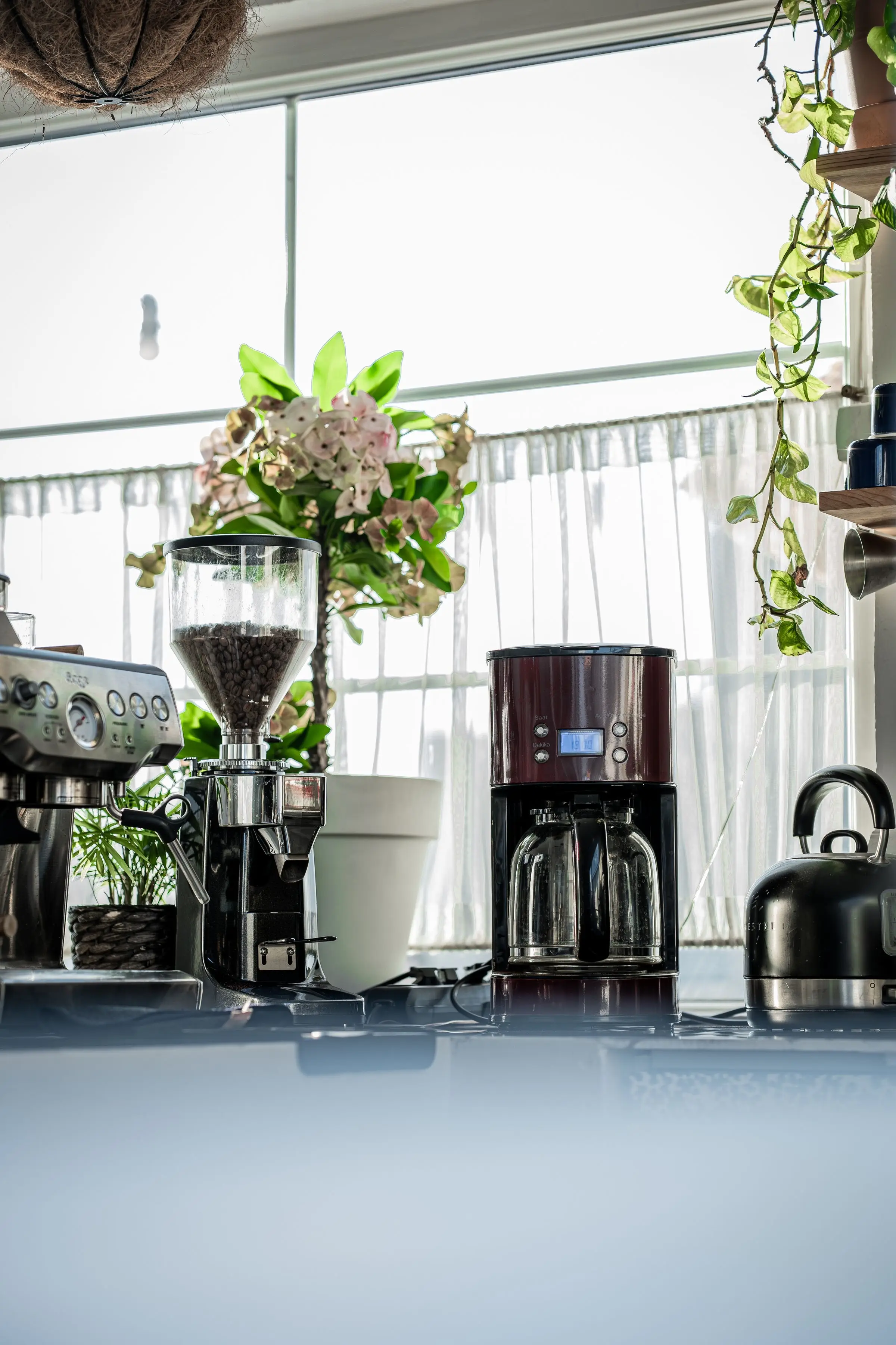 A kitchen windowsill with a grinder full of beans, a filter coffee machine with two glass jugs and a stovetop kettle.