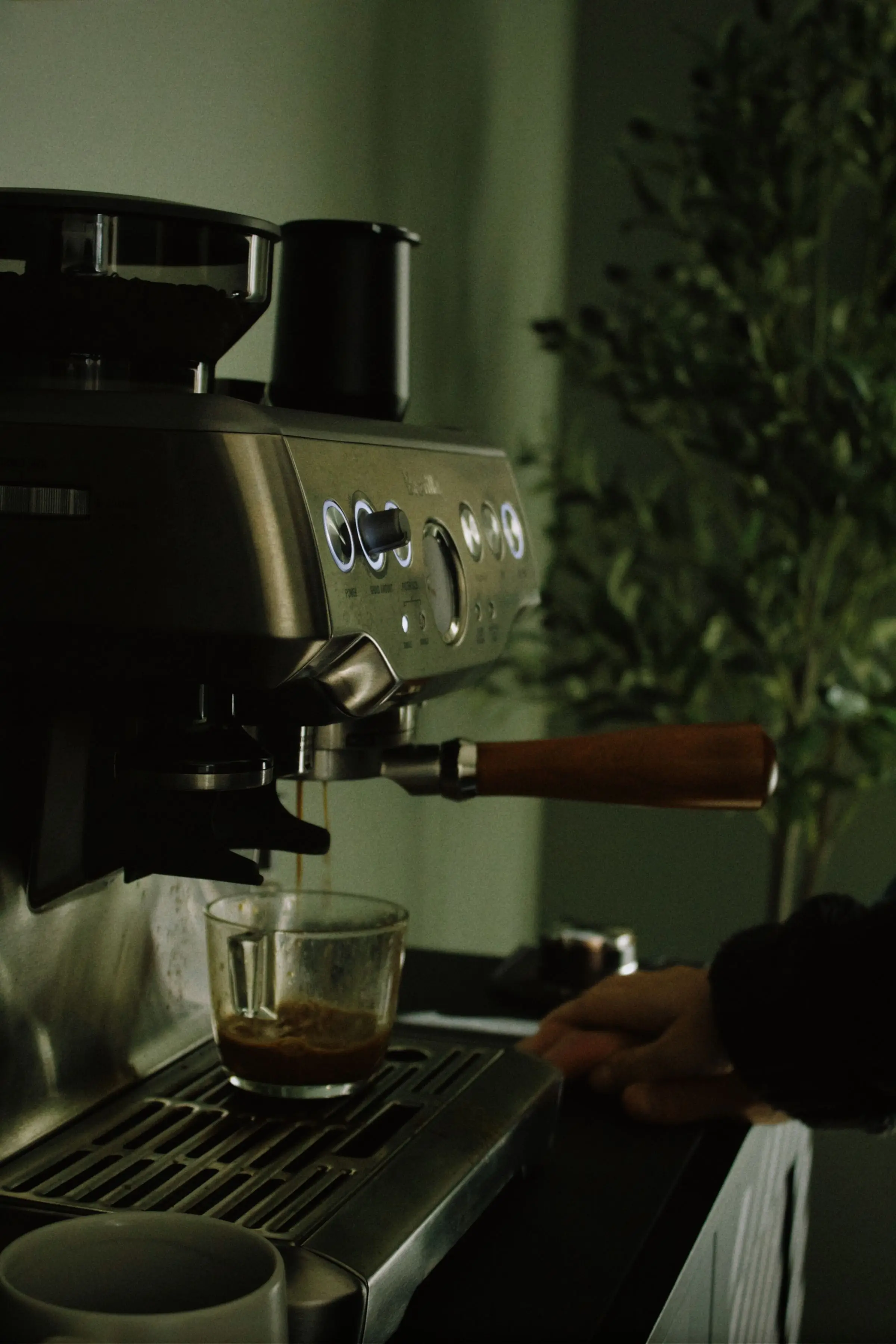 An espresso machine with a bean hopper on top and a wooden portafilter handle, filling a glass with coffee against a green wall.