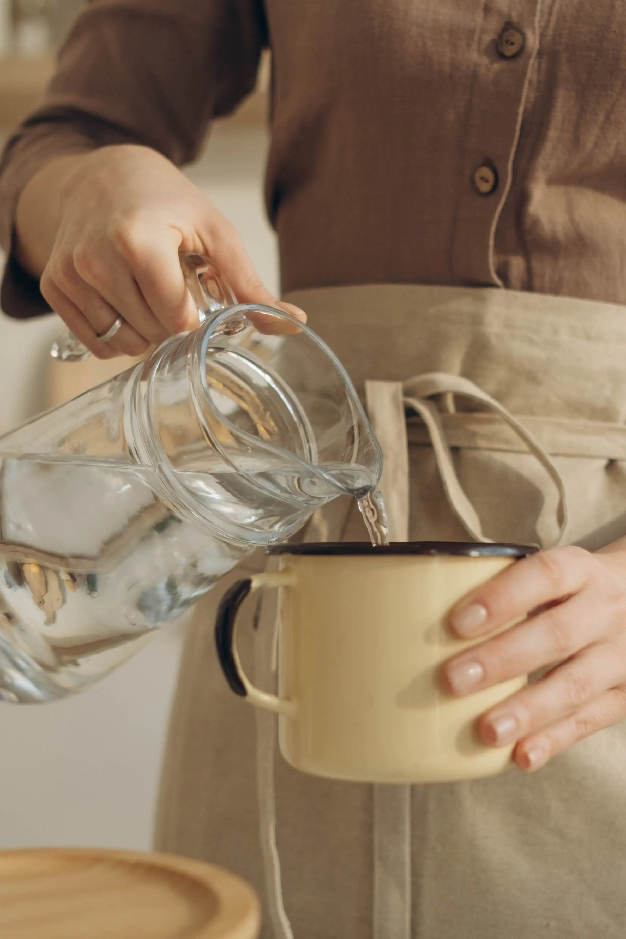 A person in a linen apron pouring water from a glass jug into a cream enamel mug.