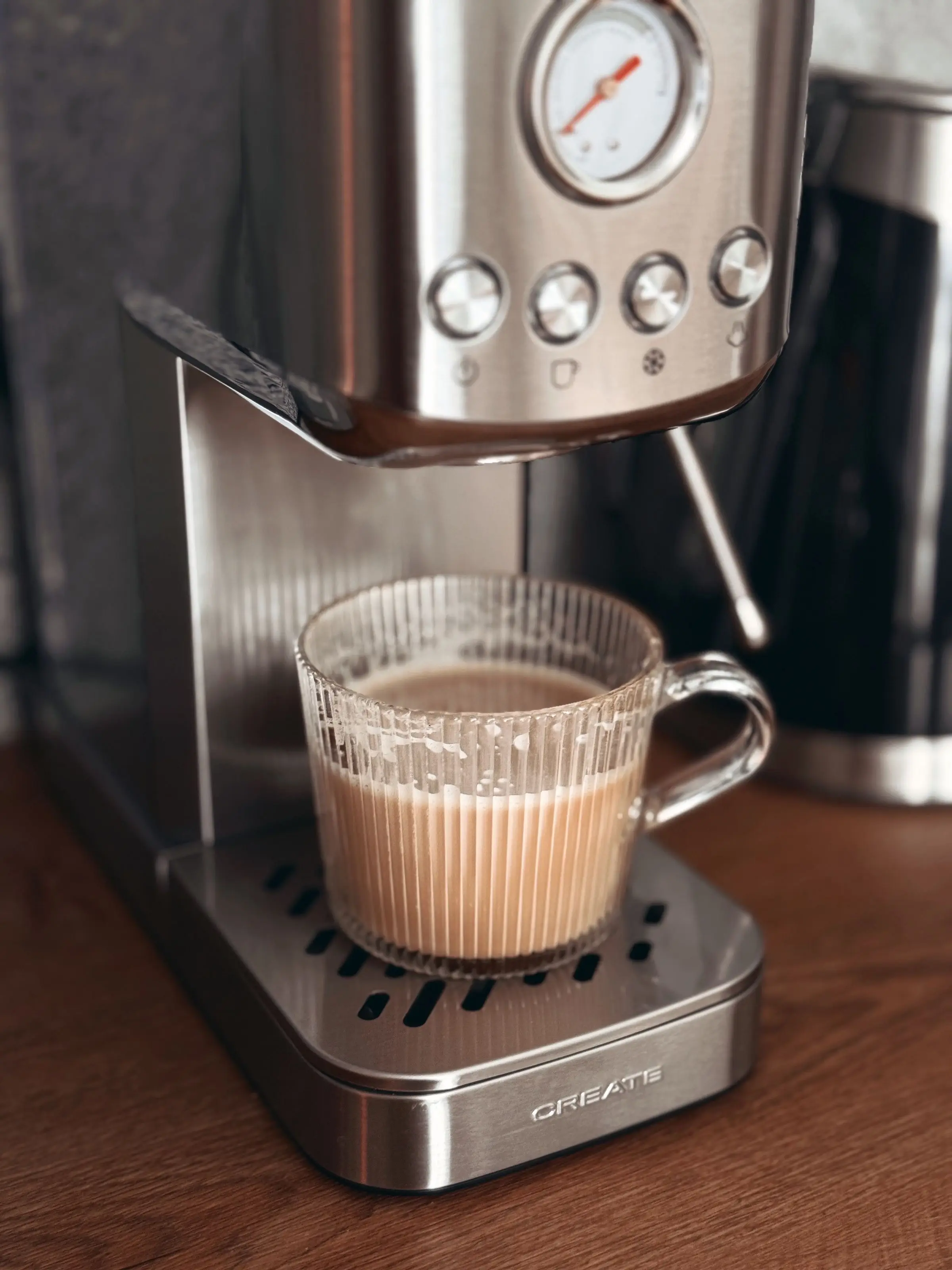 A small stainless steel espresso machine with a pressure gauge and four buttons, filling a ribbed glass cup with milky coffee on a wooden counter.