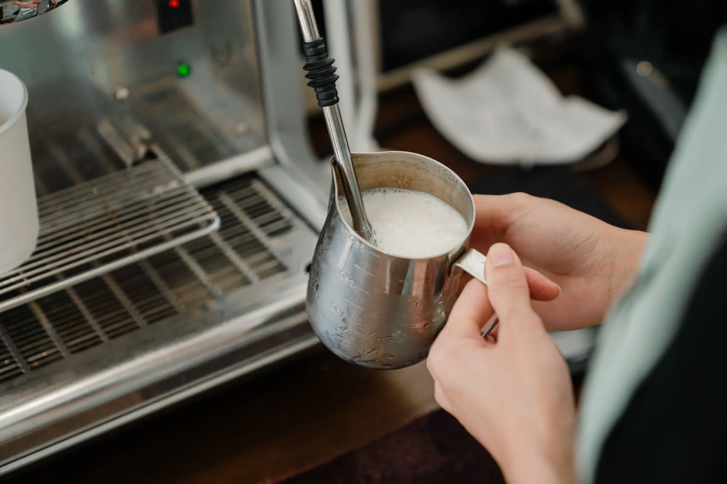 A hand holding a stainless steel jug of frothed milk under the steam wand of a coffee machine.