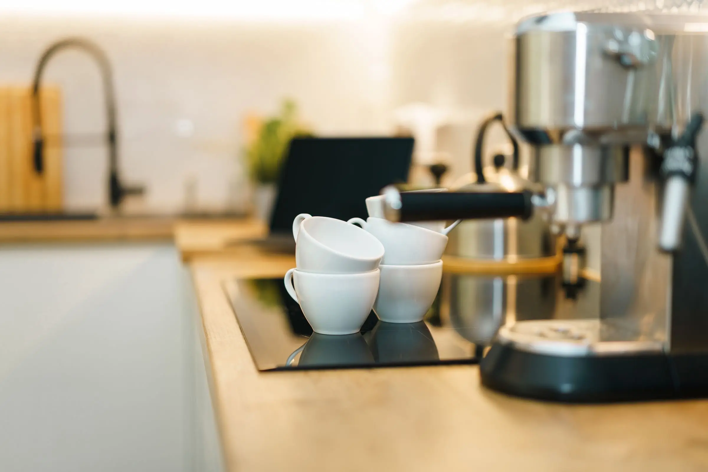 Four small white cups stacked on the warming tray of a coffee machine on a wooden kitchen counter, a black tap behind.