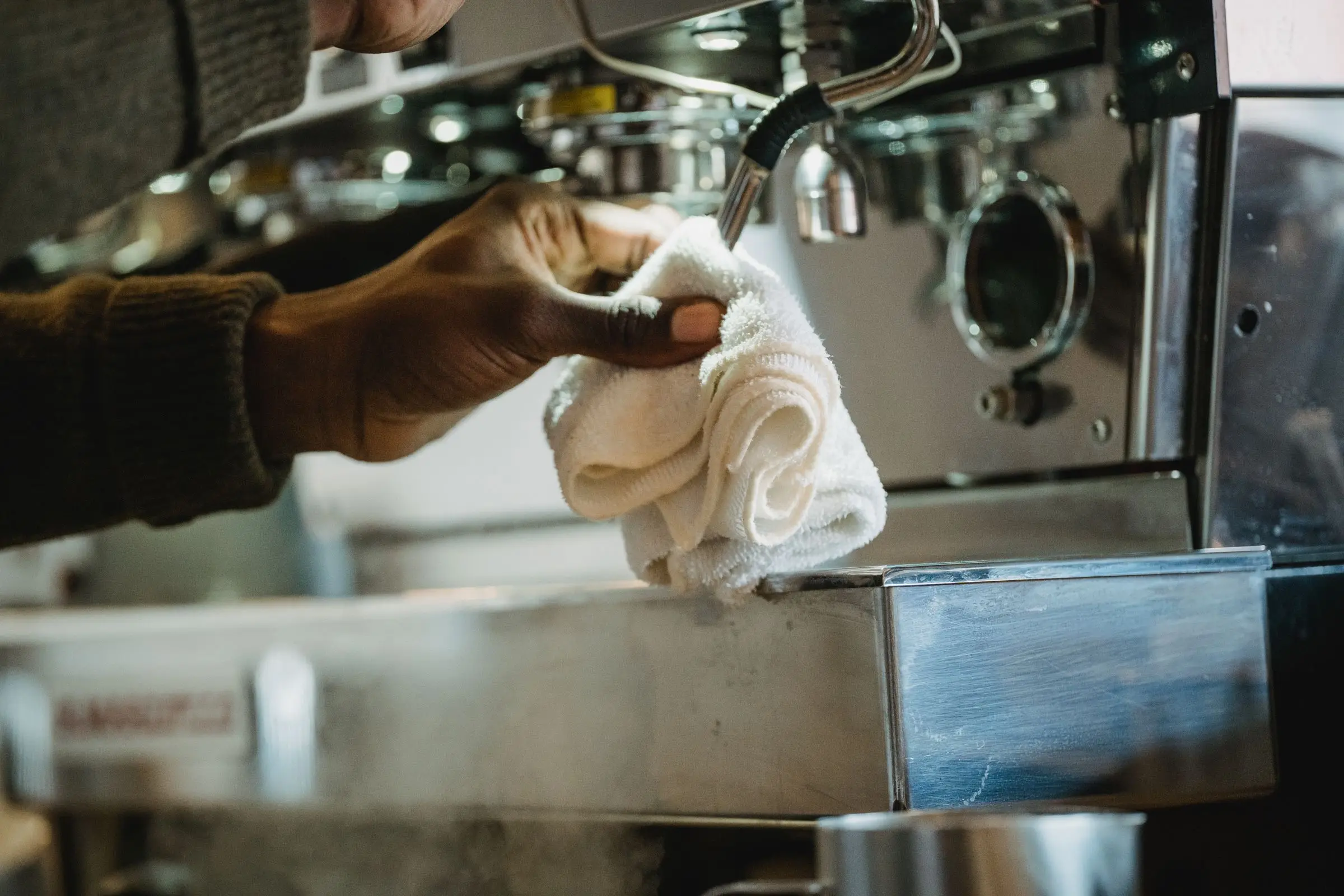 A hand wiping the front of a stainless steel coffee machine with a white cloth, steam rising from the drip tray.