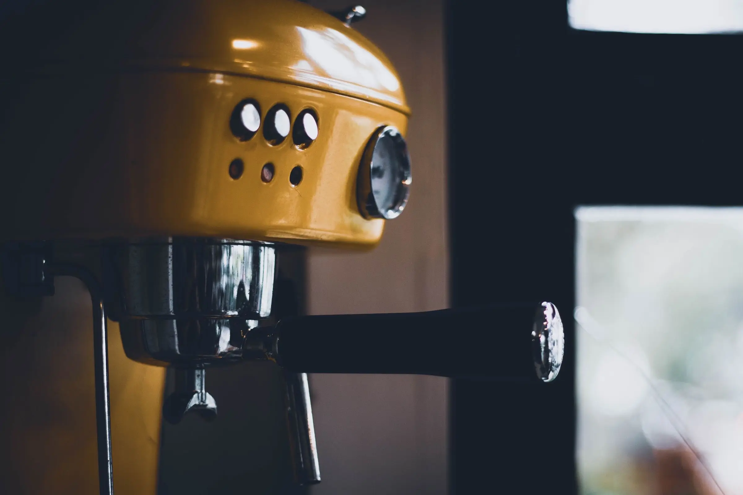 Close-up of a small yellow espresso machine, its group head and steam wand in shadow against a dark room.